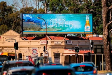 Outdoor LED billboard displaying beverage advertisement above a busy roadway with traffic and retail buildings in the background