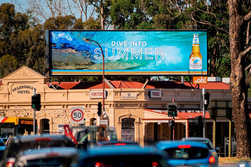 Outdoor LED billboard displaying beverage advertisement above a busy roadway with traffic and retail buildings in the background